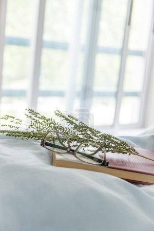 Buch und Lesebrille auf dem Bett, Fenster im verschwommenen Hintergrund. Ruhige, gemütliche und friedliche Umgebung