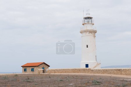 White lighthouse and small house with orange roof under cloudy sky as a background