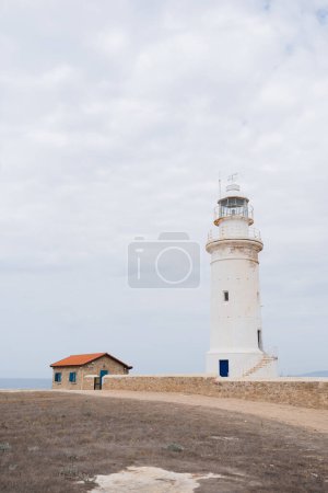 White lighthouse and small house with orange roof under cloudy sky as a background
