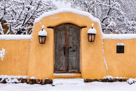 Foto de Invierno en Santa Fe, Nuevo México - muro de adobe cubierto de nieve con puerta de madera rústica y luces brillantes - Imagen libre de derechos