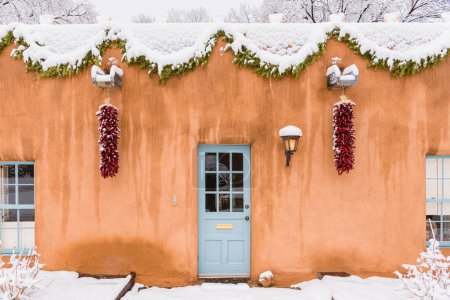 Téléchargez les photos : Scène de Noël à Santa Fe, Nouveau-Mexique, bâtiment en adobe enneigé avec ristras au chili rouge, guirlande verte et lumière éclatante - en image libre de droit