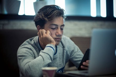adolescent seul à thome sur le canapé avec son ordinateur portable travailler ou jouer ou regarder des vidéos - nuit avec café et lunettes sur la table 