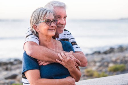 couple of seniors hogging at the beach with a lot of love - retired together - woman with glasses and man with sea background 