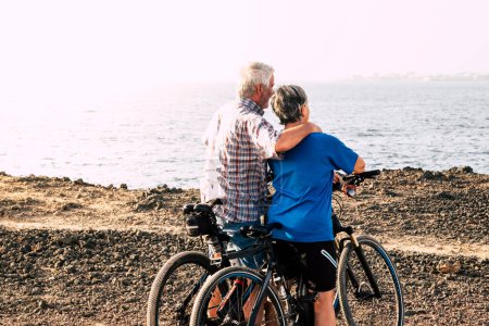 schöne Senioren, die sich am Strand oder im Park mit ihren Fahrrädern umarmten - reife und aktive Menschen, die trainieren und hart arbeiten, um fit zu sein - Meer im Hintergrund