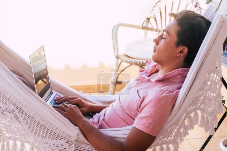 Téléchargez les photos : Adolescent seul sur la terrasse sur l'hamac regarder des vidéos, travailler ou jouer à des jeux vidéo avec son téléphone ou ordinateur et son téléphone mode de vie en ligne et les générations futures - en image libre de droit
