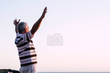 Glücklich über den Mann, der allein mit erhobenen Armen bei schönem Sonnenaufgang am Sonnenuntergang steht. Mit der Natur genießen - Rentner fühlt sich wohl und hat Spaß - Seniorenfreiheitskonzept  