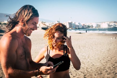 Photo for Couple of adults at the beach talking and looking at the phone of the woman stand up on the sand - woman in bikini looking at her phone and a man looking at the same phone - Royalty Free Image