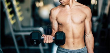 Foto de Fisicoculturista solo entrenando sus músculos en el gimnasio en silencio y sin camiseta mirando su brazo sosteniendo unas mancuernas - Imagen libre de derechos