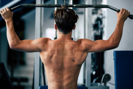 Photo for Teenager without t-shirt at the gym doing exercise for his back - man training hard with weights to be a fitness and healthy man lifestyle - Royalty Free Image