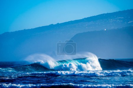 beautiful and bug wave is breaking on the sea or ocean - blue water and pacific or Atlantic - houses and mountains at the background - great place to surf 