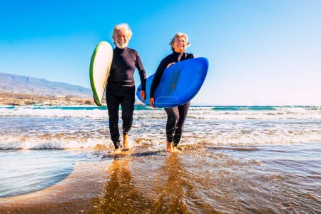 zwei ältere und erwachsene Menschen, die Spaß haben und ihren Urlaub im Freien am Strand genießen, in Neoprenanzügen und mit einem Surfbrett im Wasser mit Wellen surfen - aktive Senioren, die lächeln und Wassersport treiben 