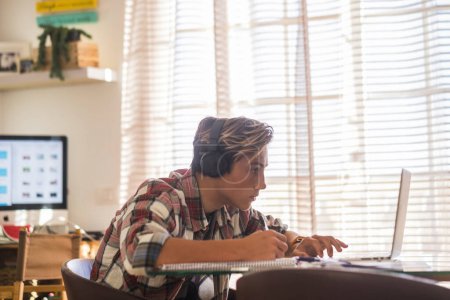 schöne Teenager Fokus für sein Studium Hausaufgaben zu Hause auf dem Tisch mit Laptop oder Computer - Kopfhörer auf dem Tisch - Indoor-Lifestyle-Konzept - Kerl schreiben und lesen 