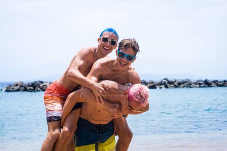 group of three friends having fun playing and enjoying together at the beach wearing sunglasses laughing smiling and looking at the camera - teenager with different colors of hair enjoying summer and vacations 