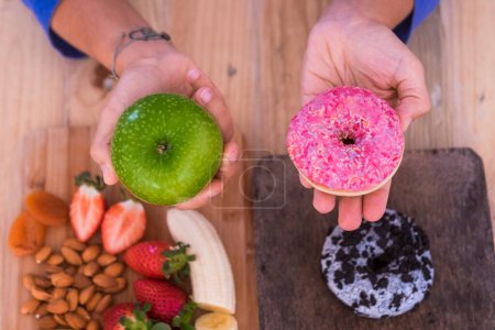 Photo for Woman choosing green apple or donut - sane and health lifestyle concept - on the table, there are one donut and some fruits - Royalty Free Image
