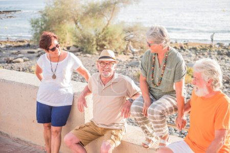Gruppe von Senioren und reifen Menschen sitzt am Strand auf einer Bank - glückliche Freundschaft mit zwei verheirateten Rentnerpaaren, die miteinander reden und Spaß haben  