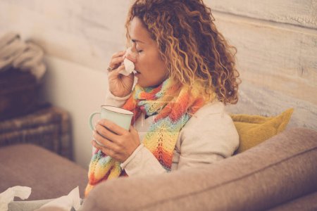 woman feeling cold, freezing, with napkin in a hand, wrapped in blanket, sitting on the sofa. Unhappy upset tired woman is suffering from a cold and using napkins - holding a cup 