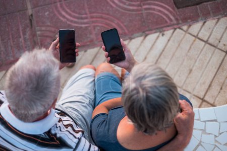 Foto de Pareja de ancianos en la playa sentados mirando sus teléfonos - pareja juntos jugando o viendo teléfonos - Imagen libre de derechos