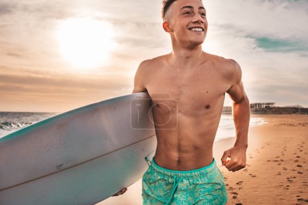 Porträt eines gutaussehenden jungen Mannes, der mit seinem Surfbrett am Strand spaziert, um in seinem Urlaub Wellen zu reiten und zu surfen 