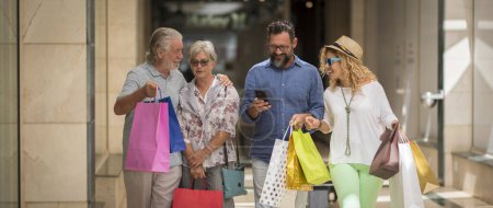 Photo for Two couples of two adults and two seniors go shopping together at the mall with a lot of bags with clothes and more on their hands - four people - man with phone - Royalty Free Image