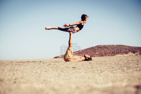 couple of two adults and people together at the beach training together on the sand doing positions of yoga with hills at the background 