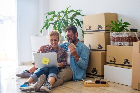 Foto de Pareja de dos adultos y personas felices después de comprar una nueva casa o apartamento juntos para vivir juntos - dos personas en el suelo usando el ordenador portátil con cajas paquetes de anuncios en la espalda - Imagen libre de derechos