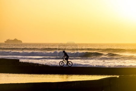 Téléchargez les photos : Adulte ou adolescent fait du vélo sur un mur du lac avec des vagues et des surfeurs en arrière-plan avec un coucher de soleil incroyable concept de liberté et style de vie - en image libre de droit