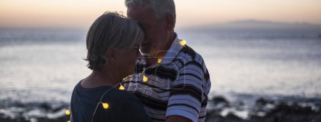 Senioren-Paar hogging am Strand mit Sonnenuntergang - Rentner-Paar mit Licht um sie herum - Liebe und Frieden Moment mit Sonnenuntergang - Ehepaar allein und romantisches Konzept - Mann umarmt seine Frau 
