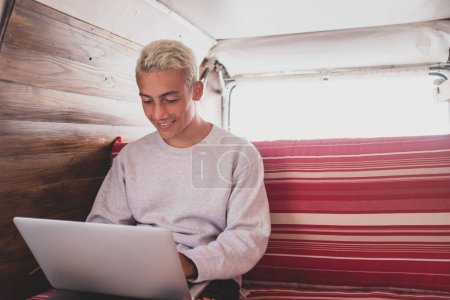 close up and portrait of young man sitting on the sofa of his minivan or vehicle - using laptop and surfing the net with social media and social network - technology nomad lifestyle 