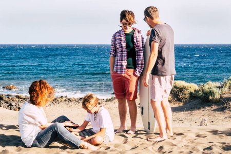 Téléchargez les photos : Famille heureuse à la plage avec un enfant et un adolescent avec planche de surf avec la mer en arrière-plan enfant et mère jouant avec le sable sur le sol - en image libre de droit