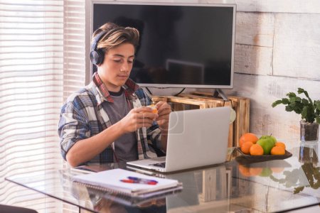 junger Teenager, der zu Hause schweigend mit Kopfhörern arbeitet - Musik hören, während er Hausaufgaben macht und eine Orange isst - Hintergrund zu Hause und viel Obst auf dem Tisch 