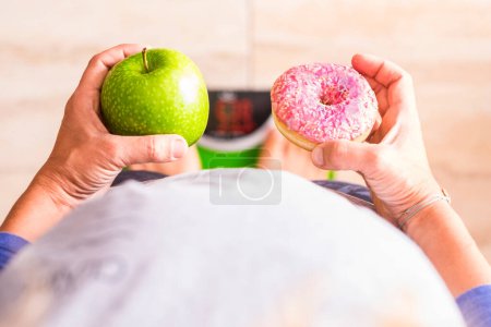 Photo for Woman look at donut and apple to select her nutrition lifestyle - she's on a weight scale - Royalty Free Image