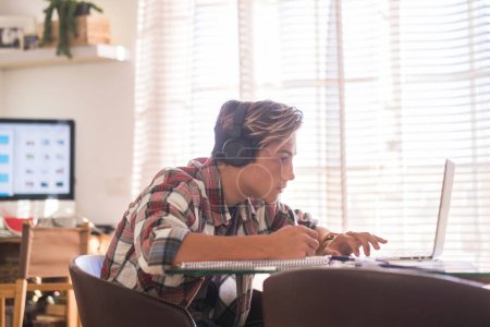 beautiful teenager focus for his studies doing homework at home on the table with laptop or computer - headphones on the table - indoor lifestyle concept - guy writing and reading  