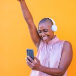 Close up and portrait of beautiful African American woman smiling and having fun dancing while listening music with headphones from her phone - enjoying using technology lifestyle   