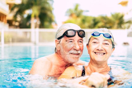 Foto de Pareja de dos ancianos abrazados en el agua de la piscina - hombre y mujer activos haciendo ejercicio juntos en la piscina - abrazados con amor - Imagen libre de derechos
