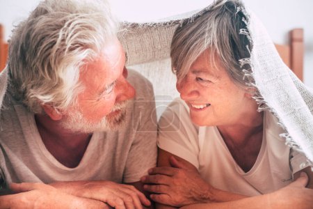 Ein paar Senioren schauen dazwischen und lächeln mit Liebe und Affekt unter der Decke am Bett - pensionierte und erwachsene Erwachsene, die morgens vor dem Aufstehen im Schlafzimmer geheiratet haben 