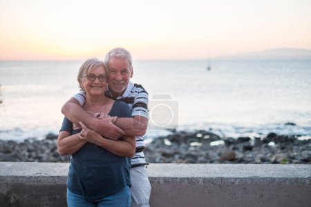 couple of seniors hogging at the beach with a lot of love - retired together - woman with glasses and man with sea background 