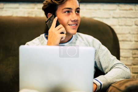teenager at home alone sitting on the sofa using his laptop while he is calling and talking at the phone - working at home or playing video games 
