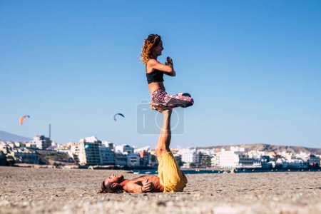 couple of two adults at the beach dong exercise like yoga called acroyoga - man lying on the sand holding a woman with his feet and leg in the air  