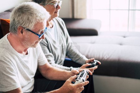 Téléchargez les photos : Couple de retraités caucasiens jouant à des jeux vidéo ensemble à la maison assis sur le canapé - femme mûre et homme marié - deux se fiancer pour toujours - tenant un contrôleur ou un joystick - en image libre de droit