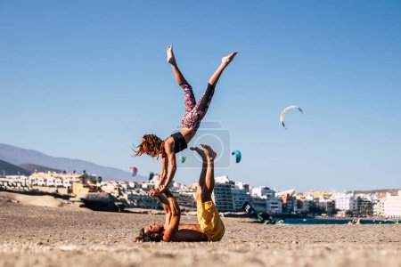 couple of two adults and people together at the beach training together on the sand doing positions of yoga with city and town at the background 