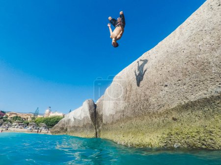Photo for Teenager jumping off on the sea by the rocks with some people looking at him - very dangerous activity and guy doing back flip and more freestyle - Royalty Free Image