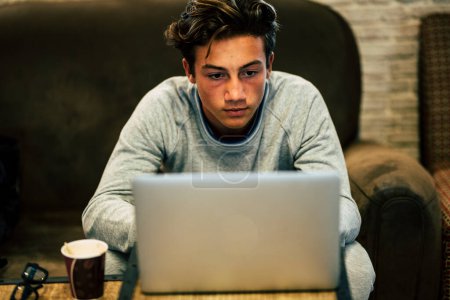 teenager alone at home on the sofa with his laptop working or playing or watching videos - night with coffee and glasses on the table 