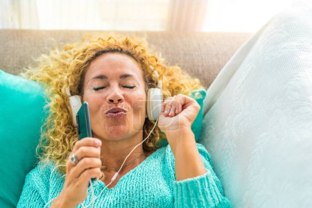 una mujer feliz y joven cantando y escuchando música en el sofá o sofá en casa con auriculares blancos - estilo de vida de musicoterapia 