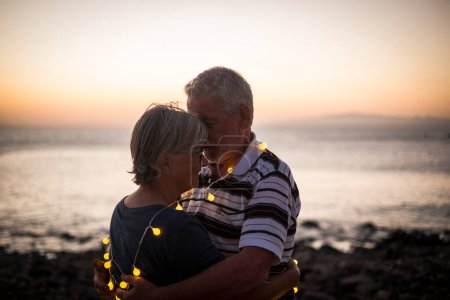 Photo for Senior couple hogging at the beach with sunset - retired couple with light around of they - love and peace moment with sunset - Royalty Free Image