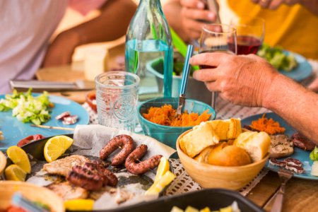 Foto de Gran mesa llena de comida vida pan, carne, verduras - algunas personas sentadas al lado de la mesa comer y beber vid y agua - celebración - un montón de ensalada y todo tipo de alimentos - Imagen libre de derechos