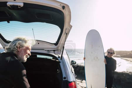 Foto de Feliz y divertida pareja de personas mayores preparando su tabla de surf en el coche para ir a la playa y probar su primera clase de surf - personas maduras activas haciendo actividades al aire libre - Imagen libre de derechos