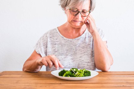 reife Frau am Tisch, die mit ekelhaftem Gesicht auf gesunde Ernährung oder Brokkoli schaut - das Gemüse anfassen und den Ernährungsstil ablehnen 