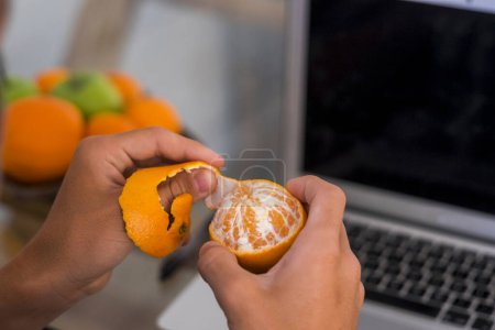Teenager beim Schälen einer Mandarine mit einem Laptop im Hintergrund und weiteren Früchten, Orangen und grünen Äpfeln 