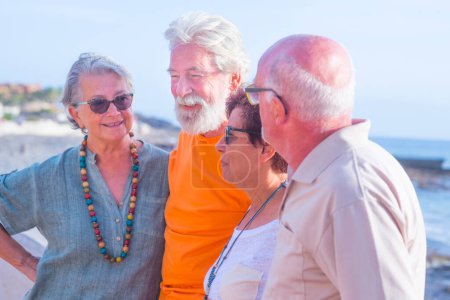 Gruppe von vier glücklichen Senioren und reifen Menschen, die miteinander reden und Spaß haben am Strand mit dem Meer im Hintergrund - Freundschafts- und Beziehungskonzept 