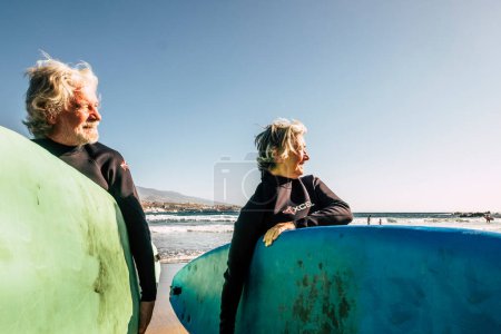 Photo for Couple of seniors at the beach with black wetsuits holding a surftables ready to go surfing a the beach - active mature and retired people doing happy activity together in their vacations or free time - Royalty Free Image